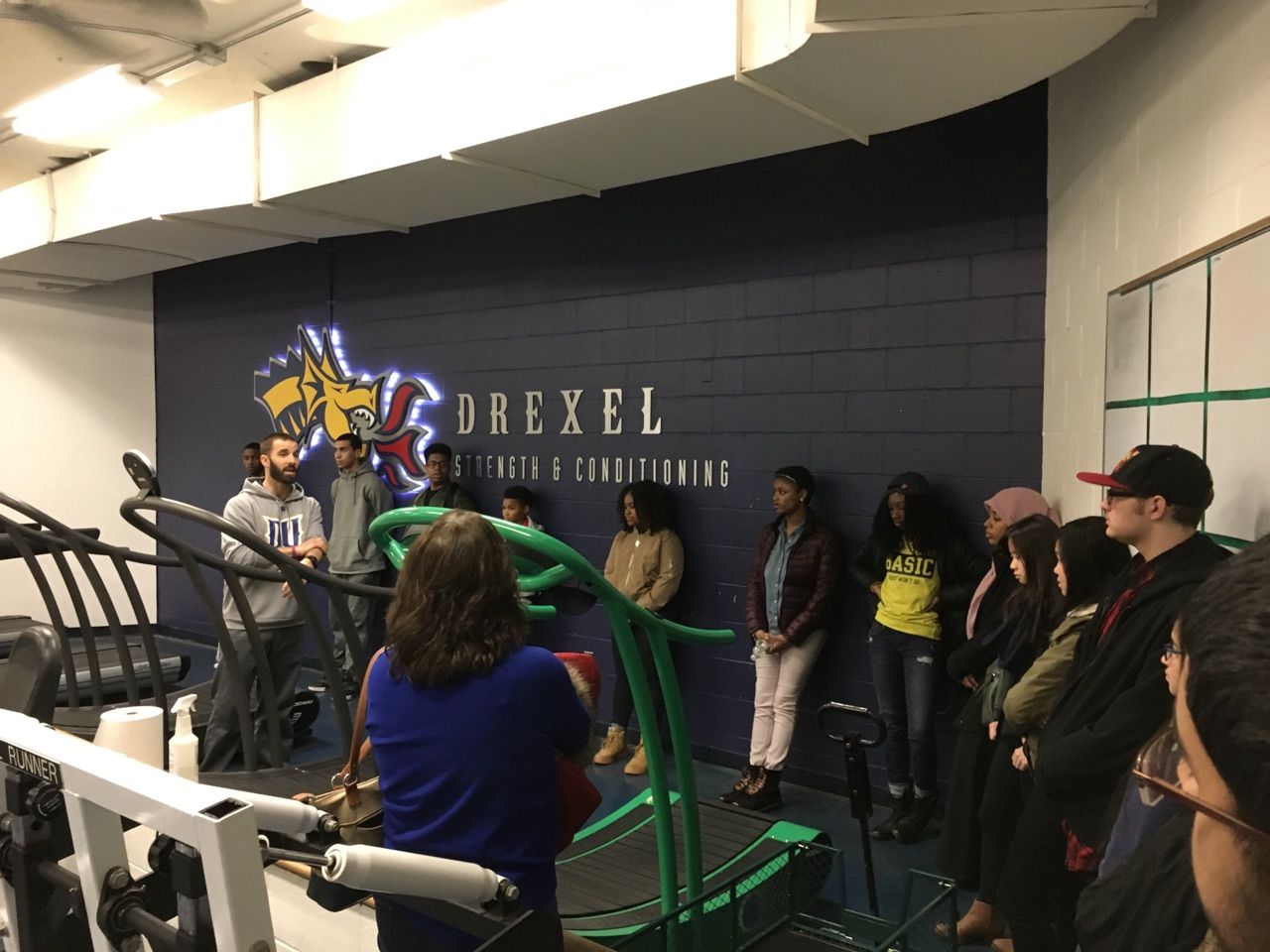 Students in the Karabots Junior Fellows Program talk with Michael Rankin, director of strength and conditioning at Drexel University. They stand in the midst of various exercise equipment (a treadmill is in the center frame).