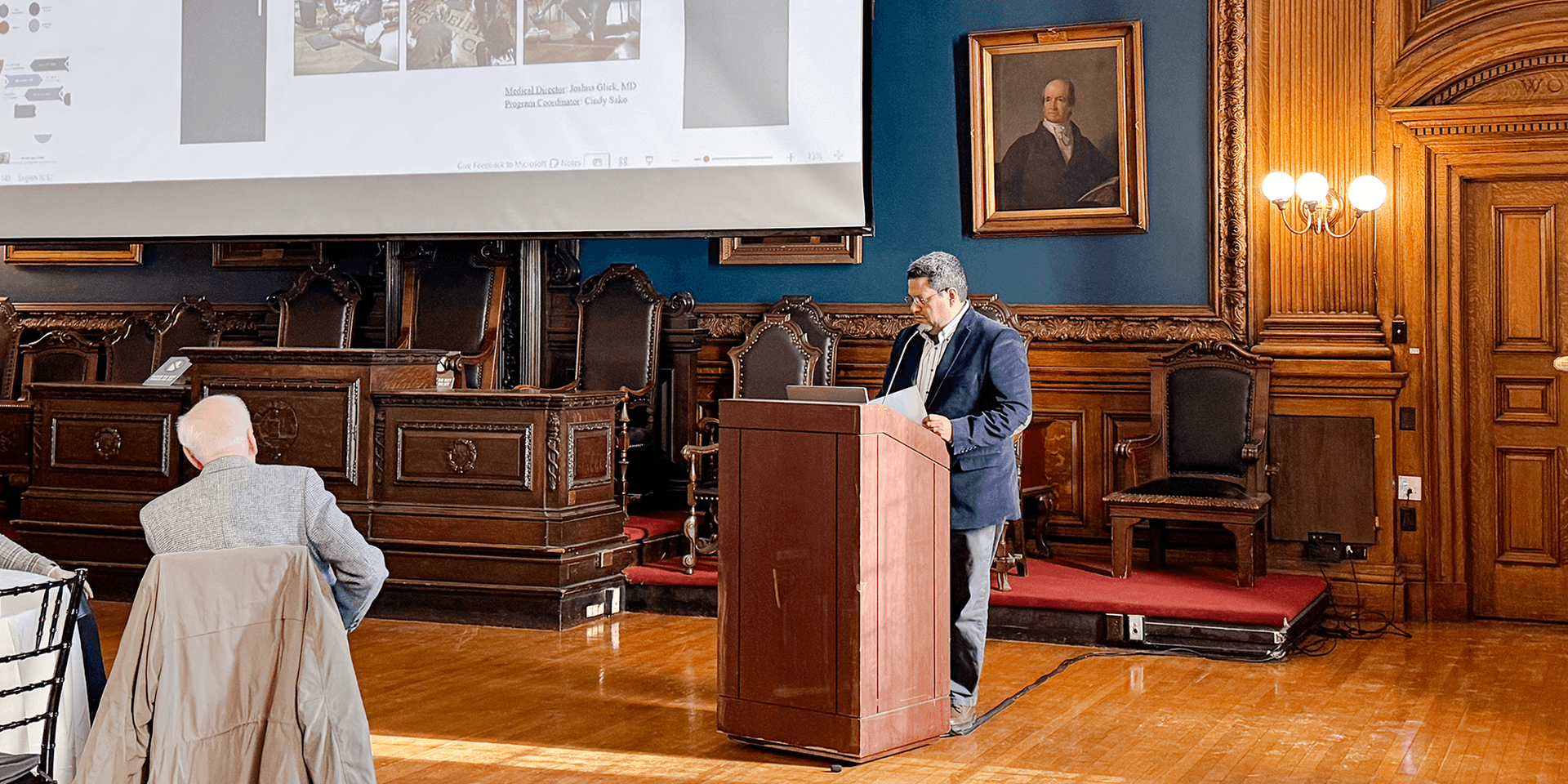 Man in dark navy suit standing at a podium in a large room with a slideshow