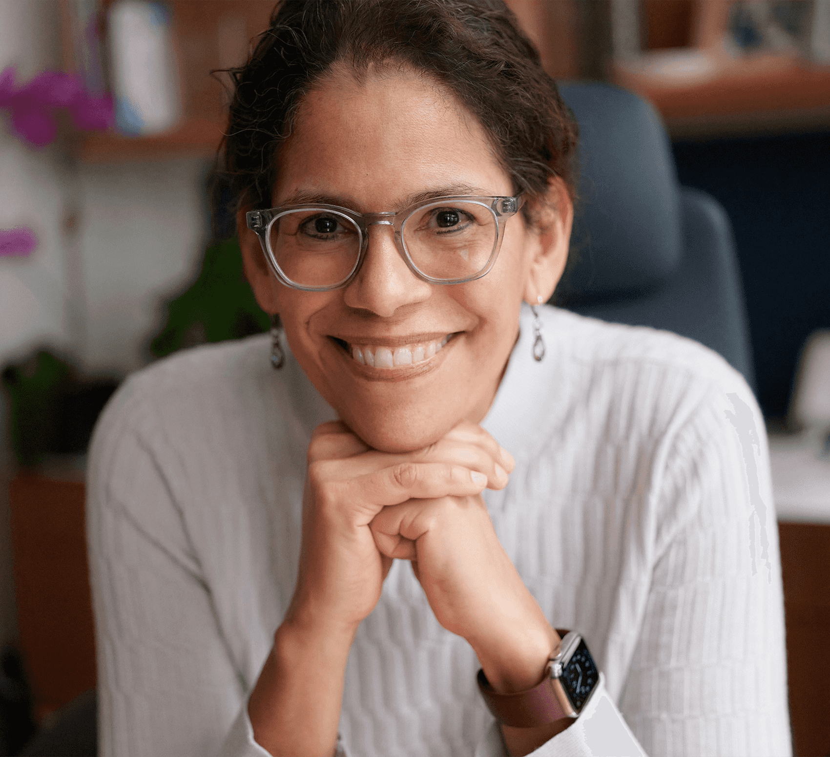 Headshot of a smiling woman with brown hair pulled back, gray glasses, dangly earrings, and a white turtleneck, resting her chin on her hands while seated at an office desk