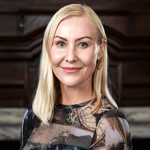Headshot of a woman with light blonde, shoulder length hair and blue eyes, in a multi-patterned dress of dark colors