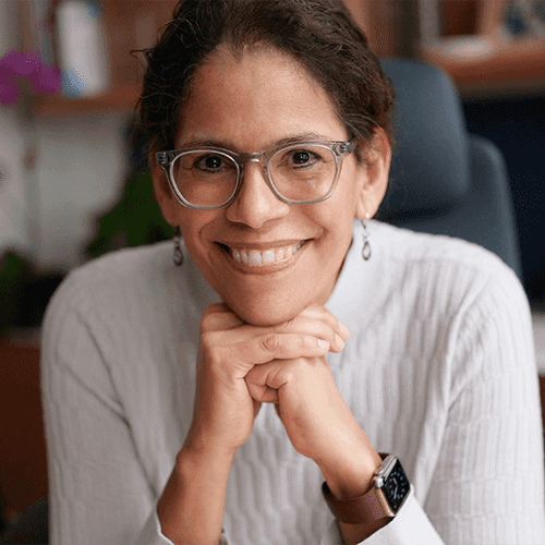Headshot of a smiling woman with brown hair pulled back, gray glasses, dangly earrings, and a white turtleneck, resting her chin on her hands while seated at an office desk