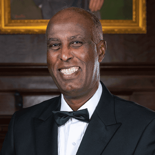 Headshot of Dr. Ibrahim standing in a black tuxedo with a bowtie against a wood paneled wall 