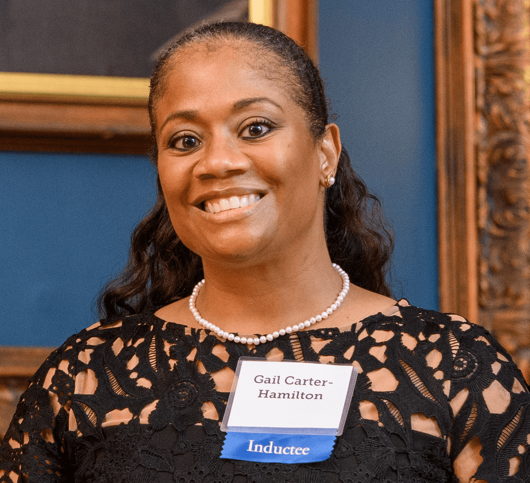 Photograph of a Black woman with dark hair pulled away from her face. She wears a black lace dress and a pearl necklace. A nametag pinned to her dress reads "Gail Carter-Hamilton" and a blue ribbon says "Inductee"
