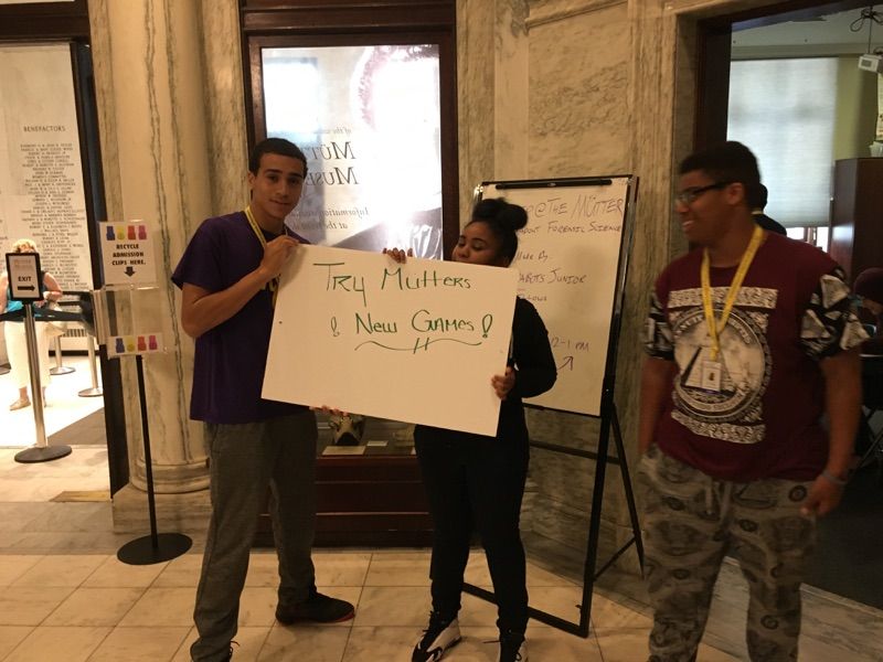 Several of the Karabots Junior Fellows entice visitors to the Müttre Museum to try their games, holding a sign saying "Try Mütter's New Games"