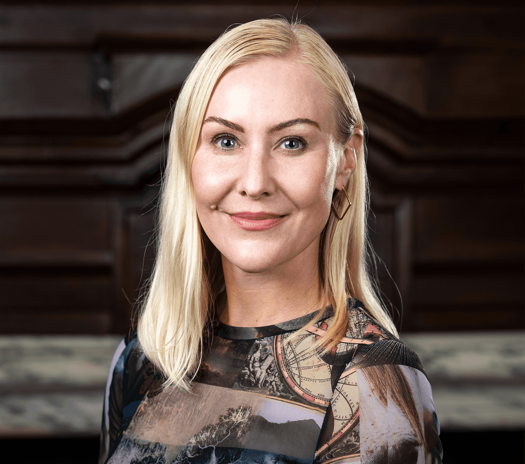 Headshot of a woman with light blonde, shoulder length hair and blue eyes, in a multi-patterned dress of dark colors