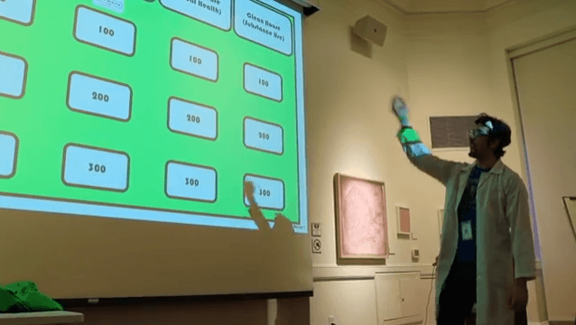 Youth Program Coordinator Kevin Impellizer (dressed in a lab coat and goggles) gestures to a projection screen, on which a Jeopardy-style game board is projected. The game took place at the "Friday the 13th @ the Mütter" event at the College of 